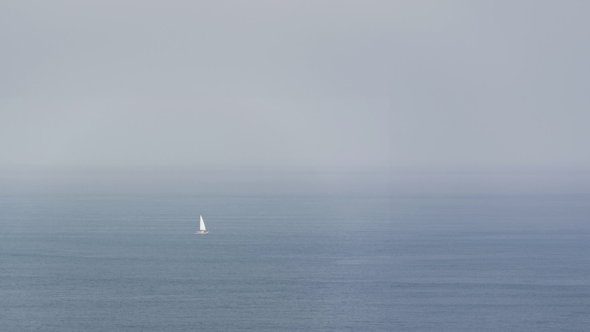A lone sailboat on a calm grey sea in fog